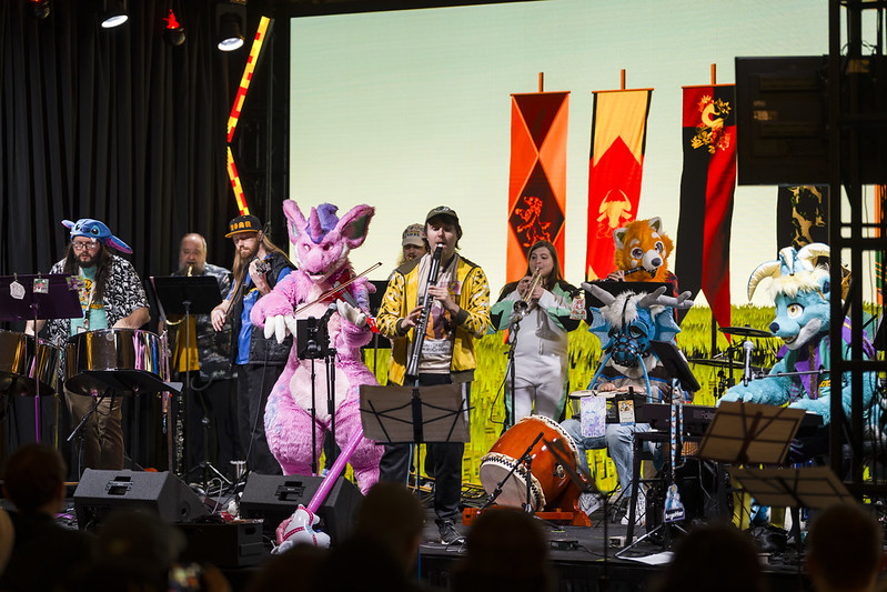 Photo of Otter Chaos! playing music on a stage. There's over 10 musicians on many different instruments. Includes a few fursuiters like Key the otter, Naoise the red panda, and Rhubarb